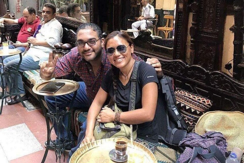A couple sitting at an outdoor cafe with a traditional wooden table and chairs, a wicker chair, and a decorative wall in the background. 5