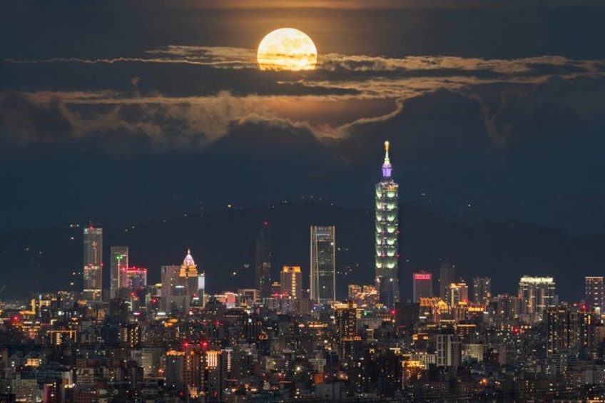 Un paisaje urbano nocturno con una torre iluminada destacada, una luna llena y un cielo nublado. 2