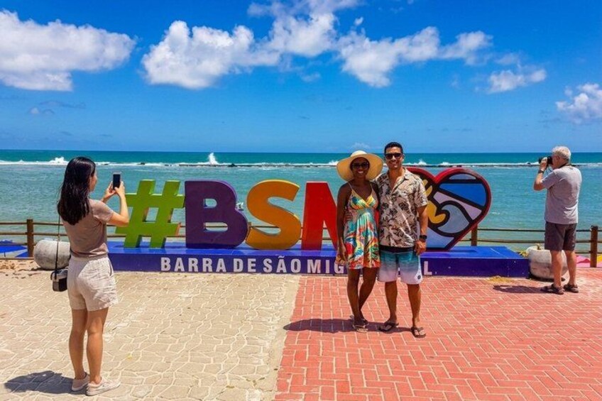 A beach scene with a sign reading 'BARRA DE SAO MIGUEL' and a hashtag '#BSM'. 2