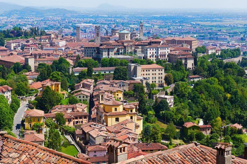 A panoramic view of a historic town with terracotta-roofed buildings, a prominent tower, and a river running through the center. 1