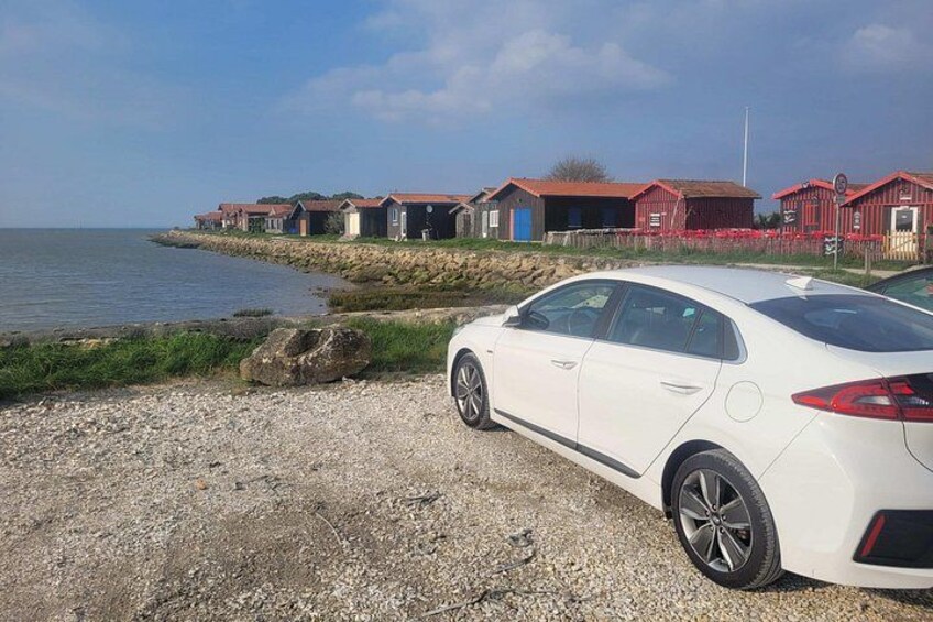 Un coche blanco estacionado en una playa de grava con edificios rojos al fondo. 2
