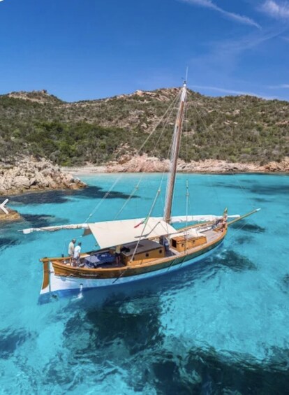 A wooden boat with a sail, anchored in clear turquoise water, with rocky terrain and greenery in the background. 3