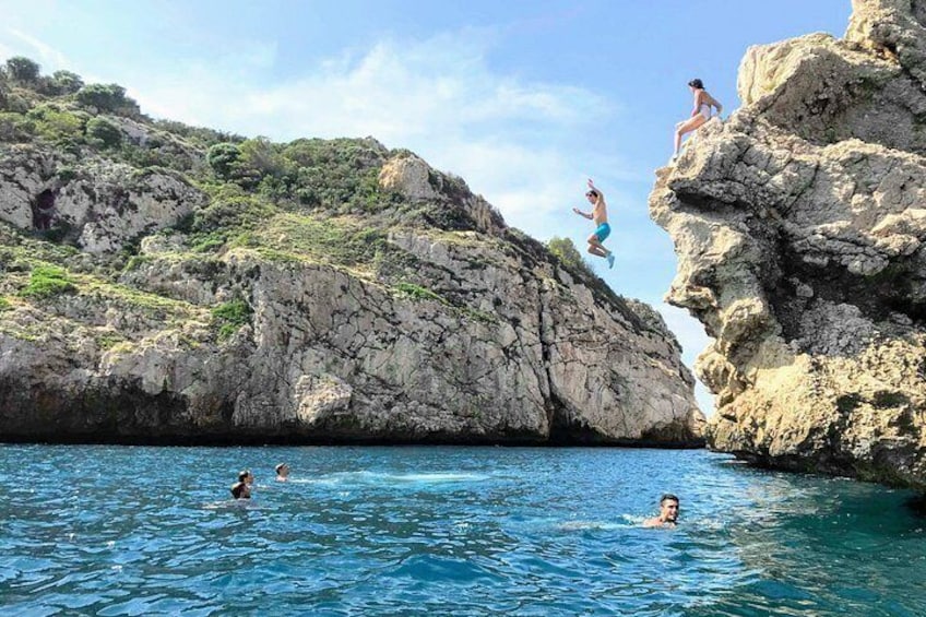 A rocky coastline with clear blue water, people swimming, and cliffs with individuals jumping off. 2