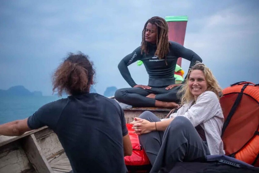 Three people sitting on a wooden platform by the sea, with one person in a black wetsuit and another in a white shirt. 4