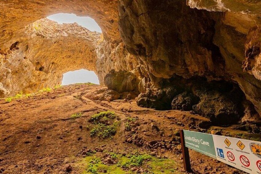 Entrada a una cueva con un sendero de tierra que se adentra, un cartel y un gran arco natural. 2