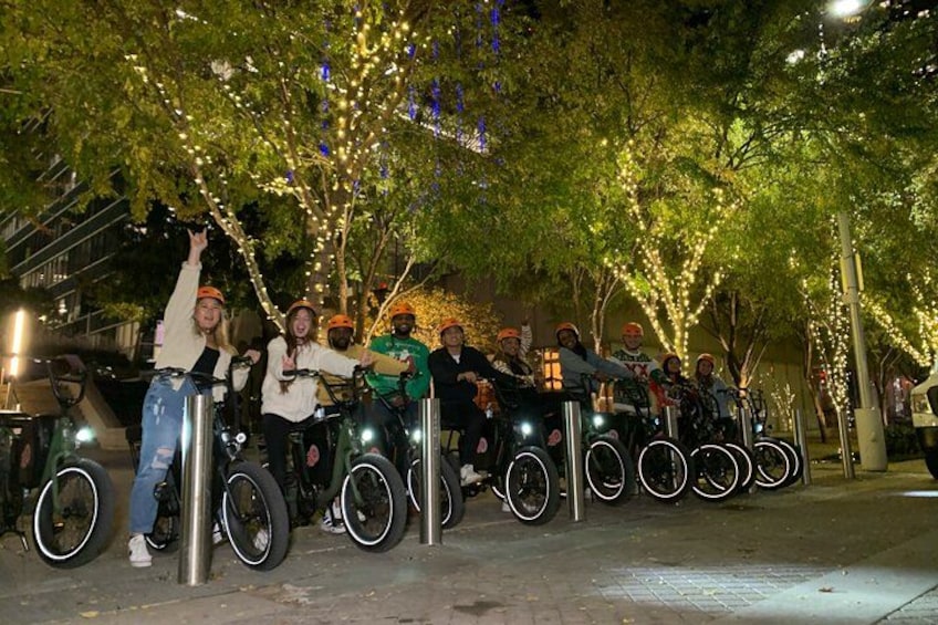 A group of people riding bicycles with large wheels, lined up along a tree-lined street at night. 1