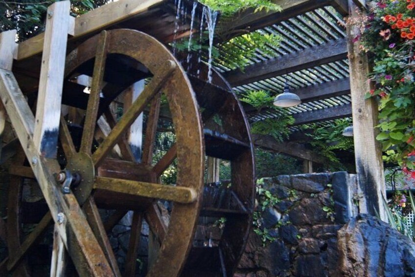 A large wooden water wheel with a wooden structure overhead, surrounded by greenery and a stone wall. 5