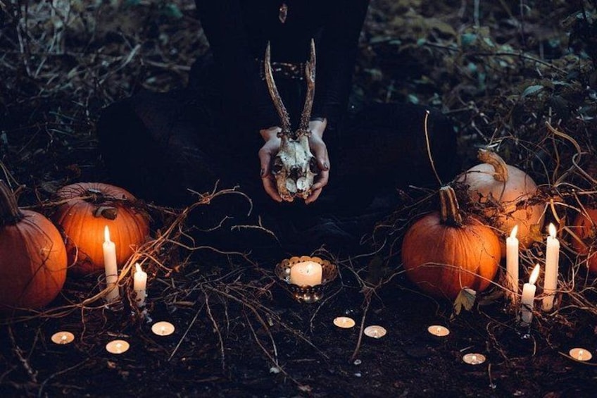 A person holding a skull with horns, surrounded by pumpkins and lit candles in a forest setting. 1