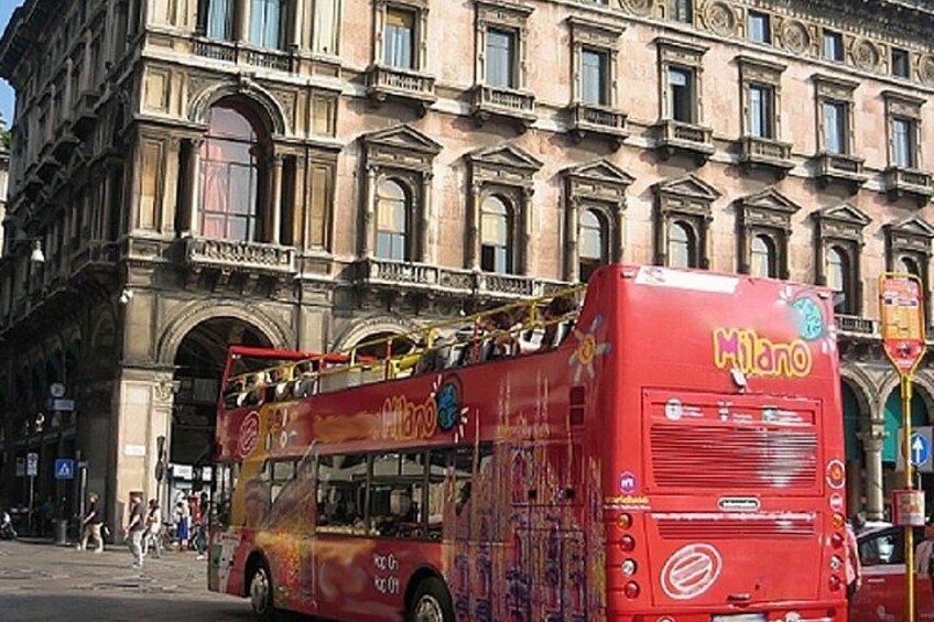 A red double-decker bus with 'Milano' written on it, parked in front of a historic building with arched windows and a balcony. 4