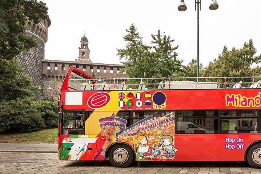 A red double-decker bus with colorful advertisements and the text 'Milanò Hop On Hop Off' parked on a street. 1