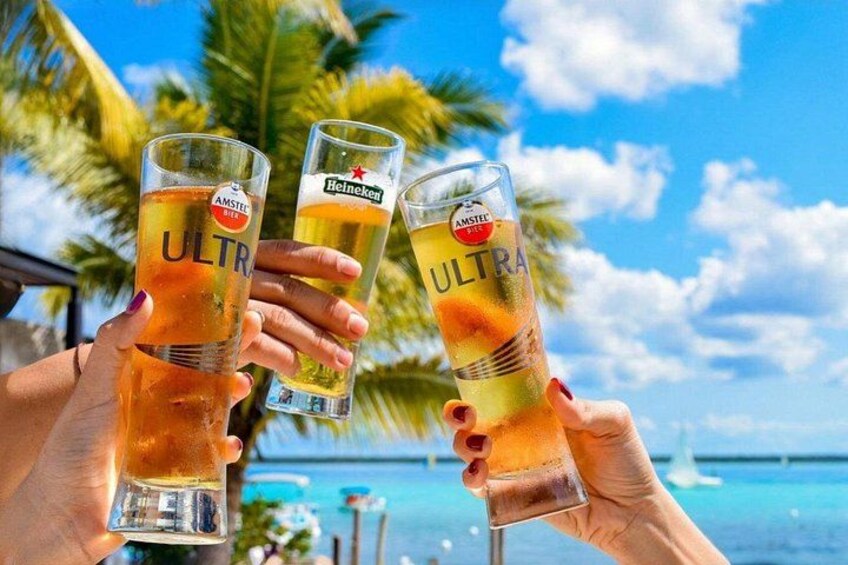 Three people holding glasses of beer with the word 'ULTRA' on them, standing by a pool with palm trees and a clear sky in the background. 3