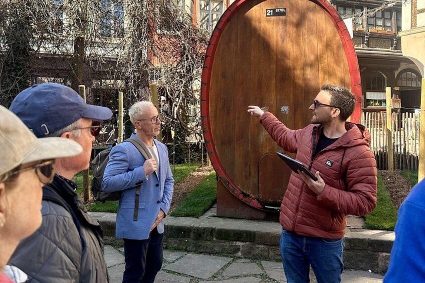 A group of people standing outdoors near a large wooden barrel with a sign above it. 2