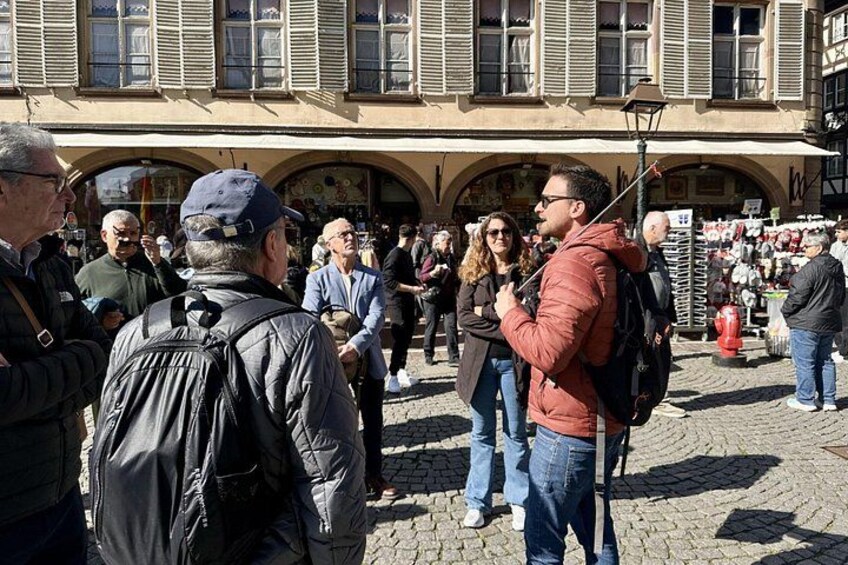 A cobblestone street with people standing and walking, a building with arched windows, and a market stall in the background. 3