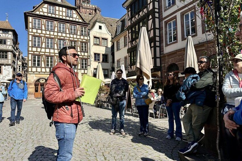 A cobblestone street with people standing and walking, historic buildings with timber framing, and outdoor seating areas. 4