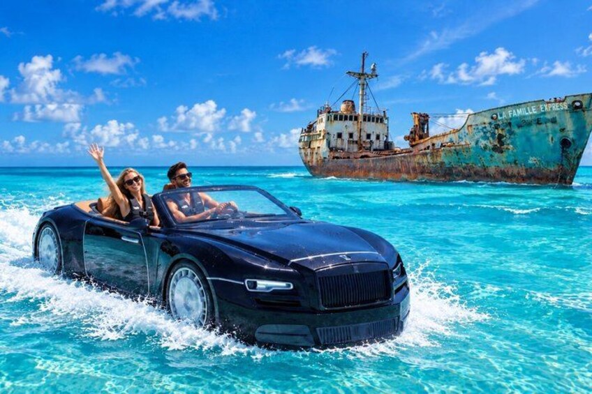 Ein Cabrio fährt durch türkisblaues Wasser, im Hintergrund ein rostiges Schiff. 1