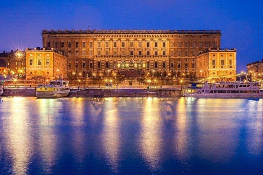 A large, illuminated building with a reflection in the water, a boat docked in front, and street lights along the waterfront. 4