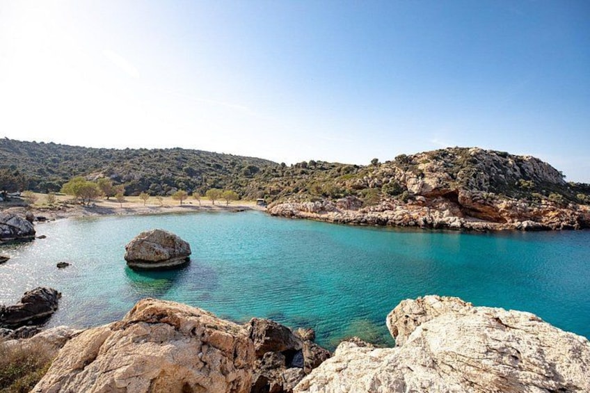 A coastal landscape with clear turquoise water, rocky outcrops, and a hilly background. 1