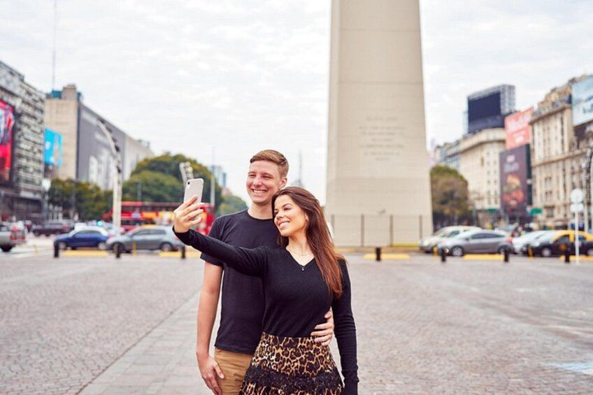 A couple taking a selfie in a city square with a tall monument in the background. 1