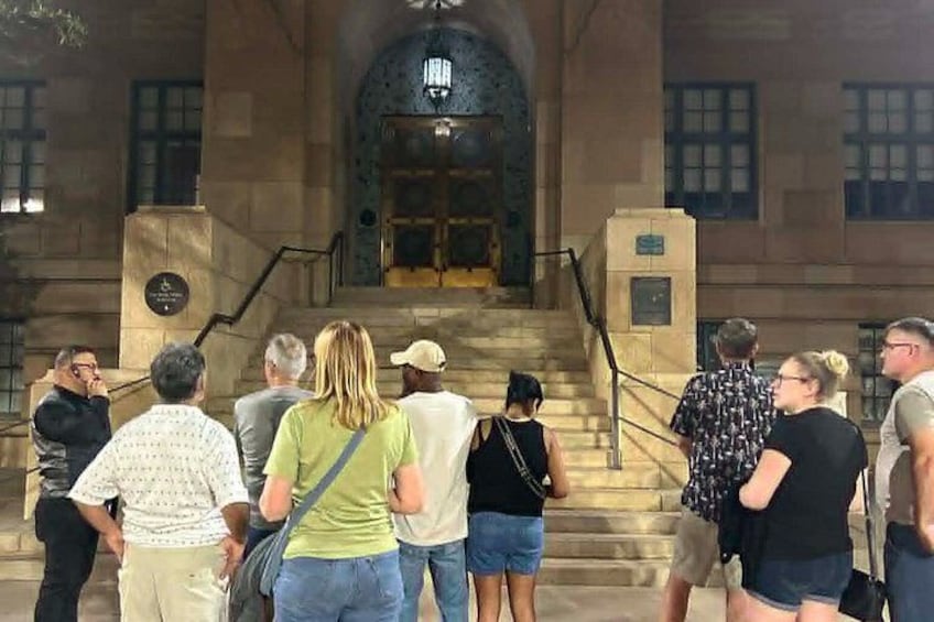 Un groupe de personnes se tient devant un bâtiment doté d’une entrée imposante et d’un escalier menant à l’intérieur. 2