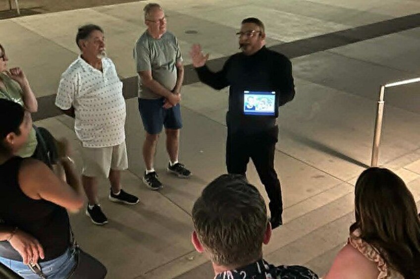 Un grupo de personas reunidas al aire libre de noche, algunas de pie y otras sentadas, con una persona sosteniendo un dispositivo que muestra texto. 4