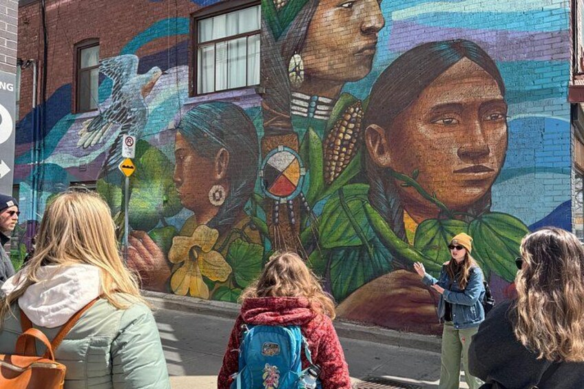 A mural with indigenous figures and nature on a brick wall. 1