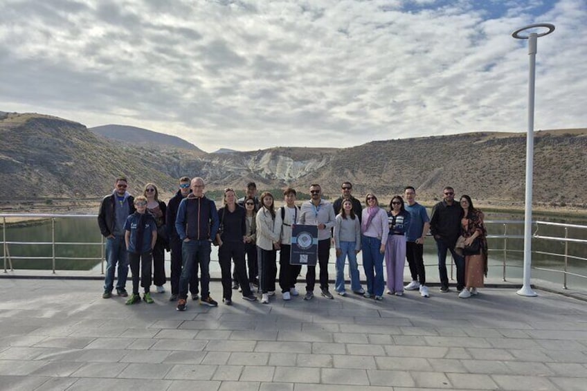 Un grupo de personas de pie en una zona pavimentada con barandilla, con vista a una masa de agua y colinas al fondo. 4