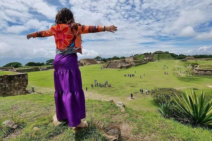 A person in a purple skirt and orange top with arms outstretched, overlooking an ancient archaeological site with ruins and green grass. 2