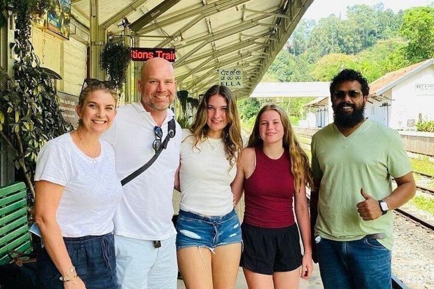 A group of people standing at a train station platform with greenery and a building in the background. 5