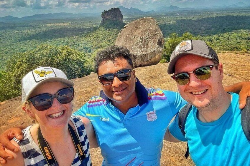 Three people standing on a hill with a view of a landscape that includes a large rock formation and greenery. 4