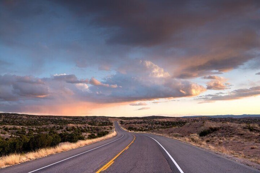 A road curving through a desert landscape with sparse vegetation and a dramatic sunset sky. 1