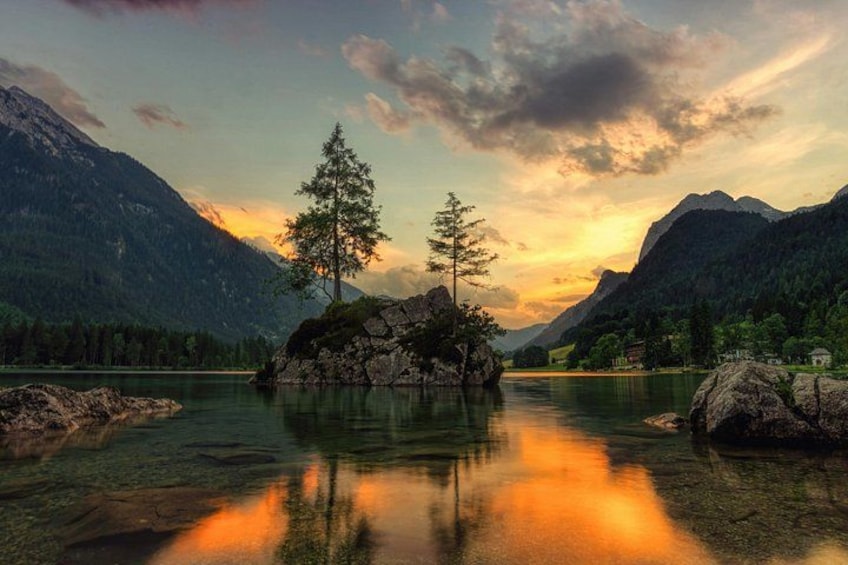 A calm lake with a reflection of a sunset, surrounded by mountains and a single tree. 3