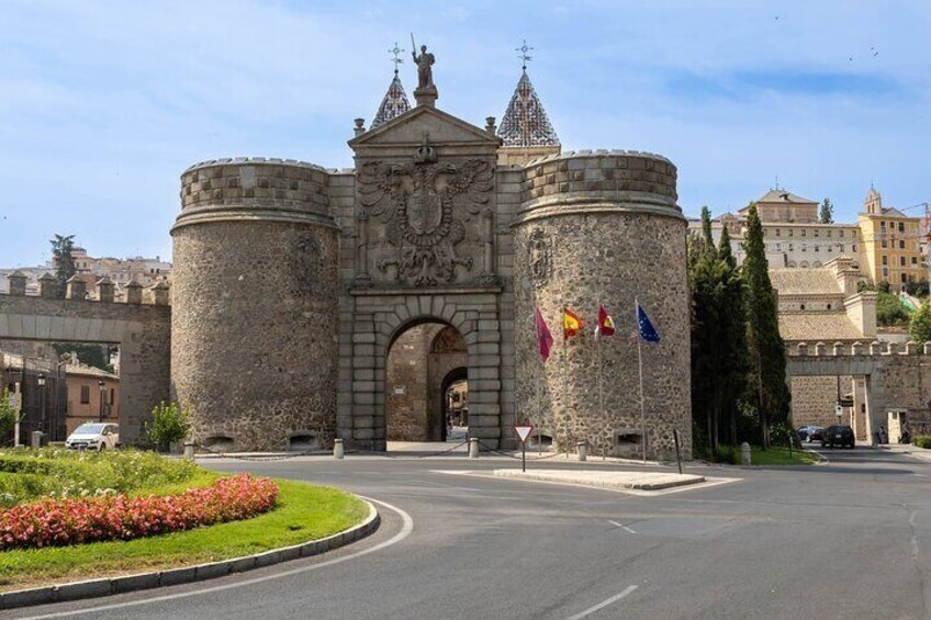 Een historische stenen poort met vlaggen, een ronde toren en een geplaveid weggetje ervoor. 1