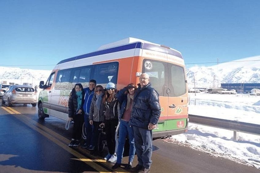 Eine Gruppe von Personen steht vor einem Bus auf einer verschneiten Straße, im Hintergrund sind Berge zu sehen. 4