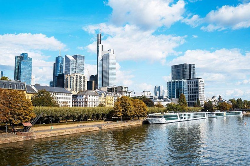 A city skyline with modern skyscrapers, a river with boats, and a park with trees. 1