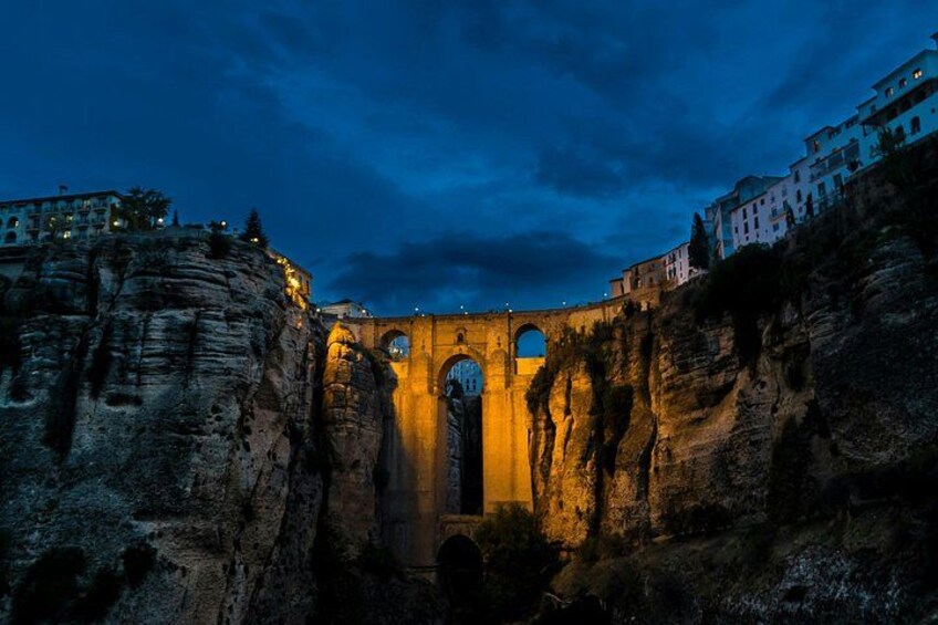 A stone bridge with arches, surrounded by rocky cliffs and illuminated buildings. 2