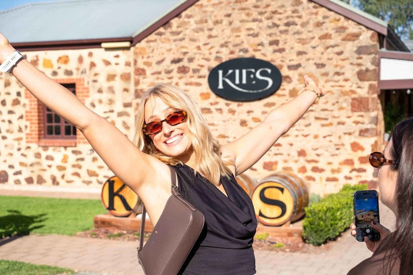 A person taking a selfie with raised arms in front of a building with a sign that reads 'KIES'. 1