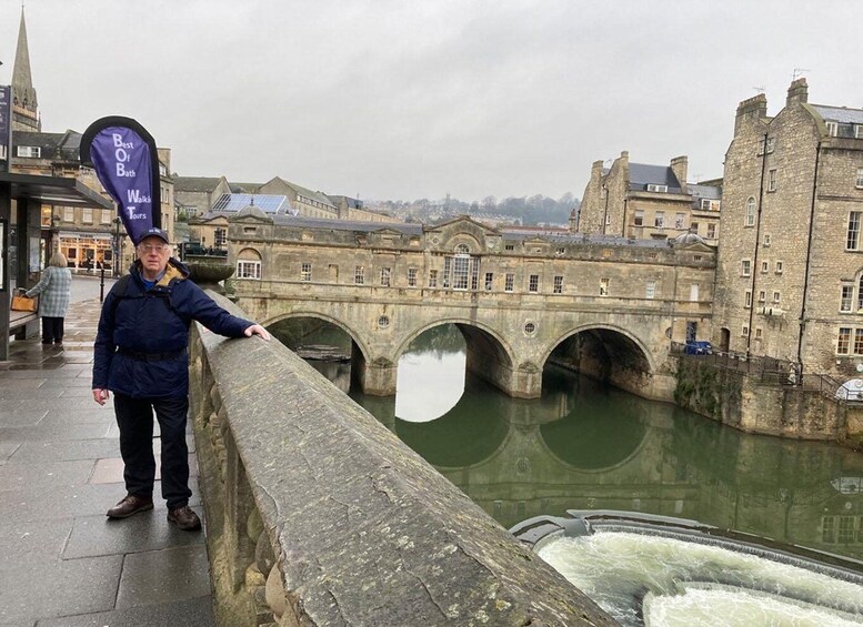 A person standing on a stone bridge with a river below, overlooking historic buildings and a cloudy sky. 1