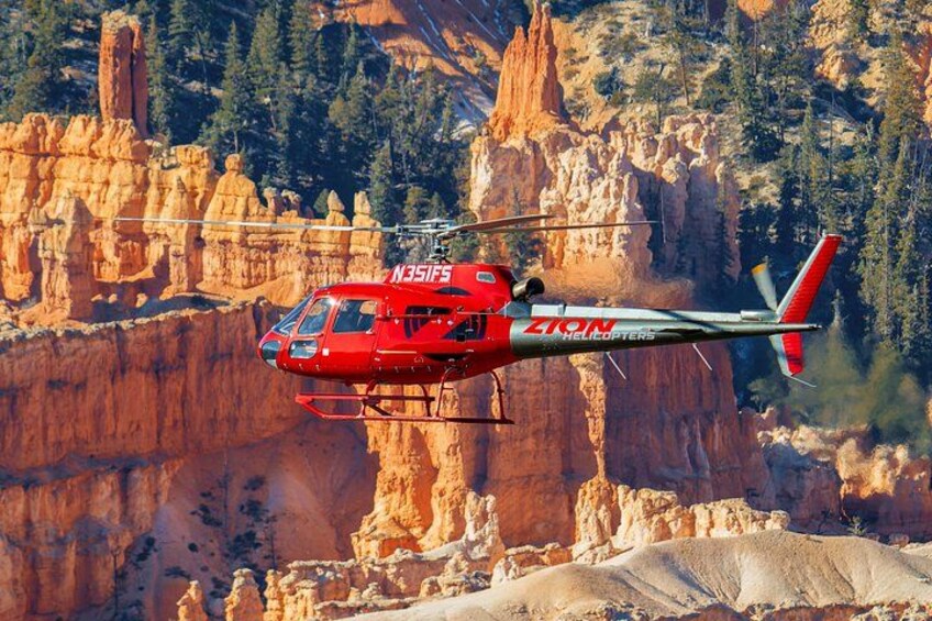 Un hélicoptère rouge, portant les inscriptions « ZION Helicopters » et « N351FS » sur son flanc, survole un paysage accidenté et rocheux où la végétation est clairsemée. 3