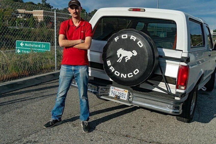 En hvid Ford Bronco med et reservehjul monteret bagtil, en mand stående ved siden af bilen, og et vejskilt der viser Mulholland Dr. 2