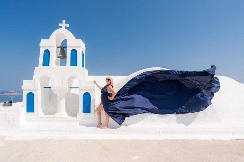 Une personne vêtue d’une robe bleue, debout devant une église blanche aux fenêtres bleues, avec une cloche. 3