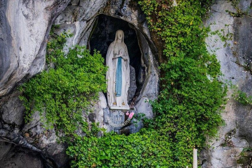 A statue of a religious figure in a cave-like setting with green foliage. 1