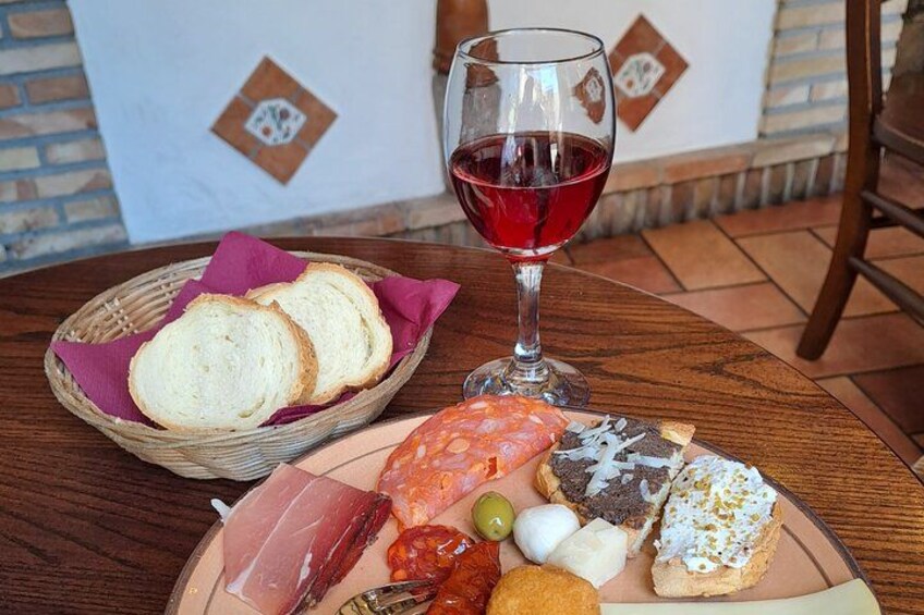 A table with a basket of bread, a glass of red wine, and a plate of assorted food including olives, cheese, and bread. 4