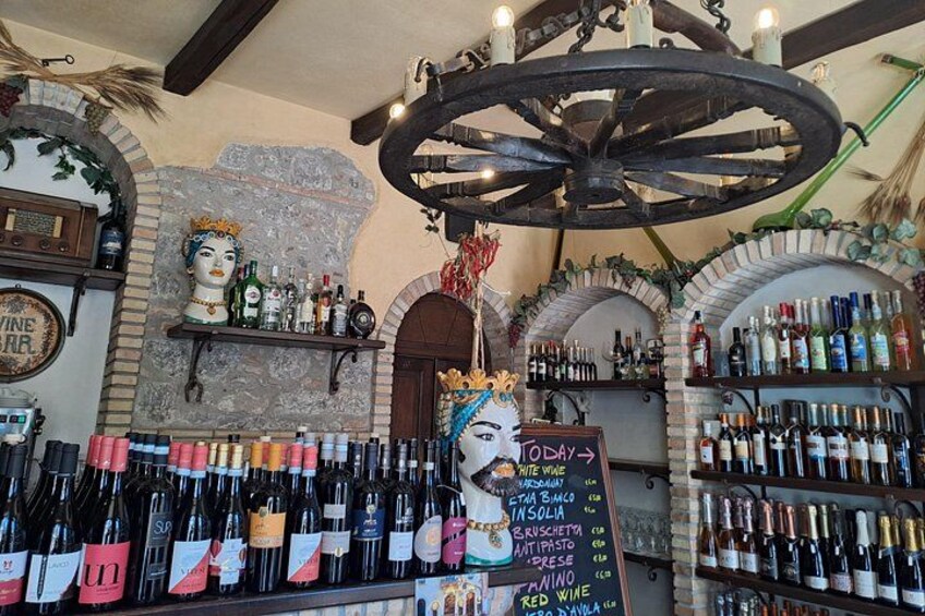 A bar with a vintage aesthetic, featuring a large wheel chandelier, a mannequin with a mustache, and shelves stocked with various bottles. 3