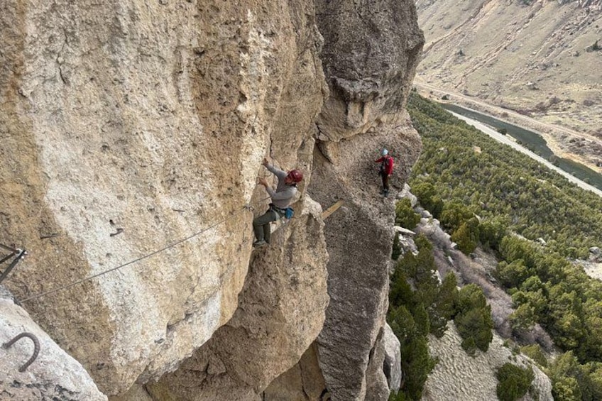 A person climbing a steep rock face with a river and forest in the background. 1