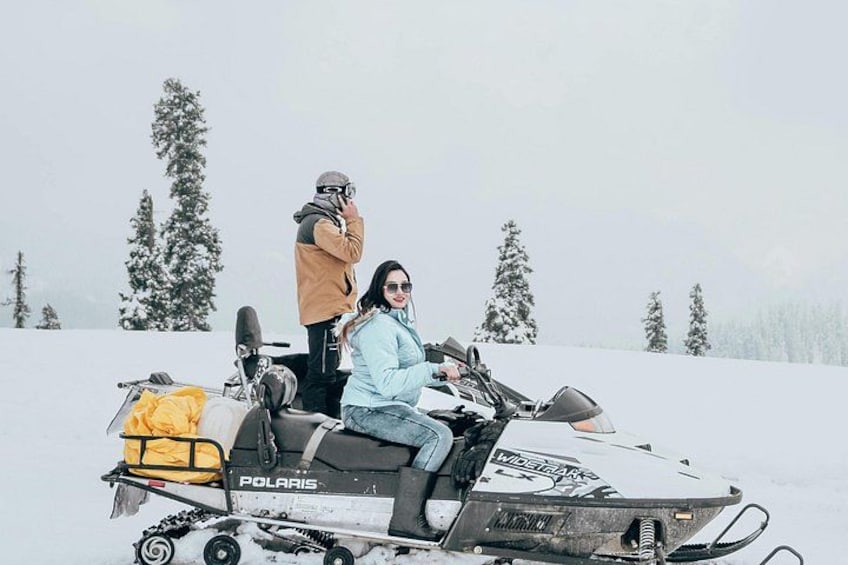 Un vehículo para nieve con una persona sentada y otra de pie a su lado, en un paisaje nevado con árboles. 2
