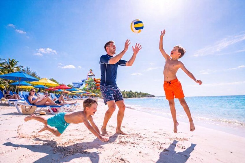 Menschen spielen Beachvolleyball an einem sandigen Strand, im Hintergrund Sonnenschirme und Palmen. 2