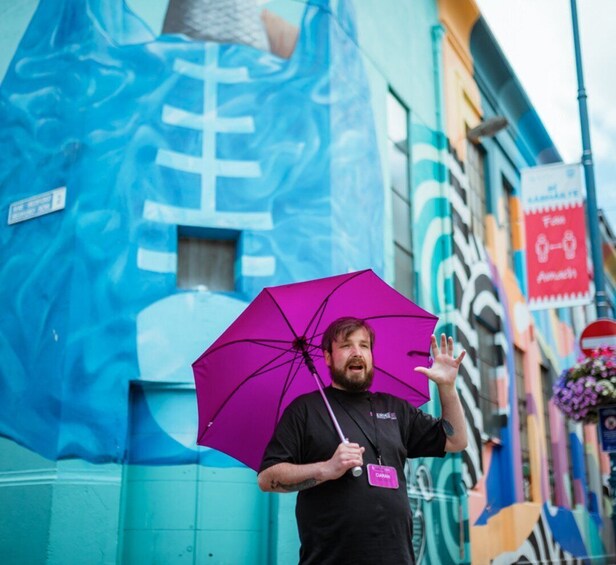 A person holding a purple umbrella, standing in front of a colorful mural on a building. 3