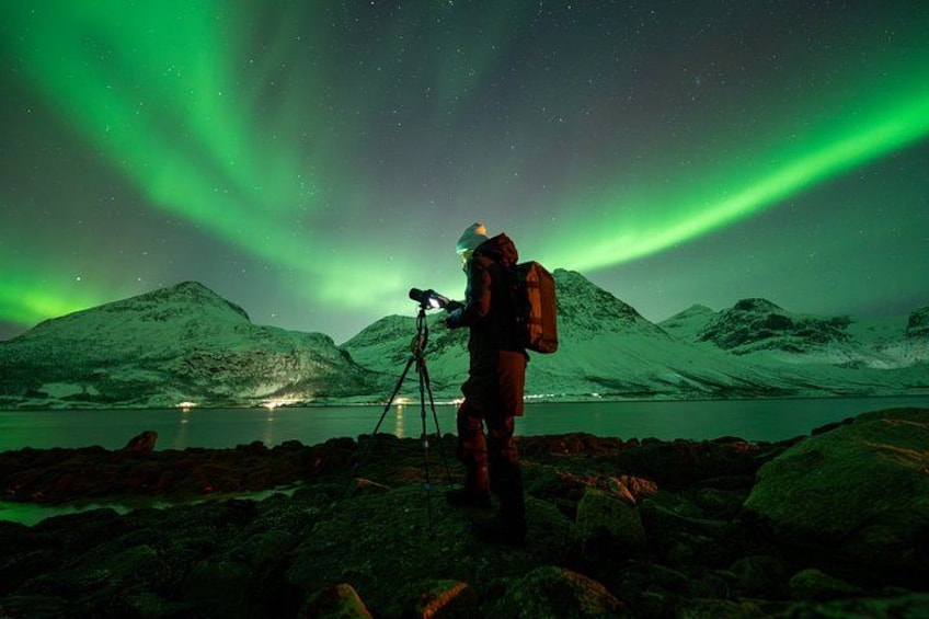 A person with a telescope and backpack, observing the Northern Lights over a mountainous landscape. 1