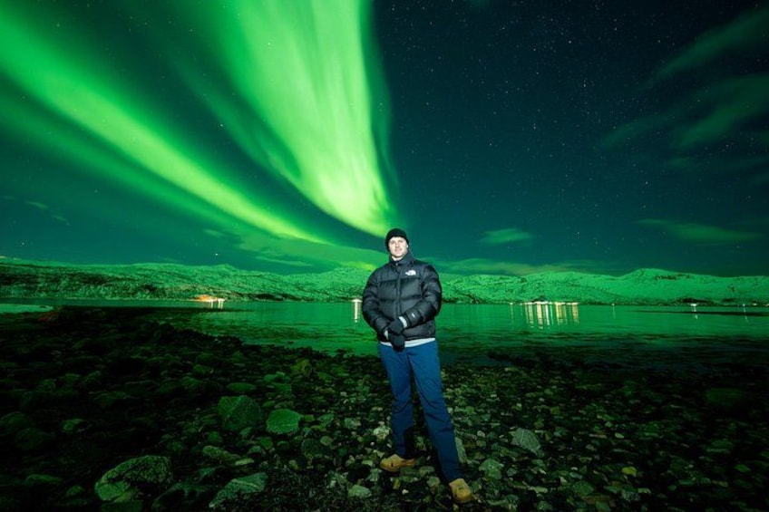 A person standing on a rocky shore with green aurora borealis in the background. 4