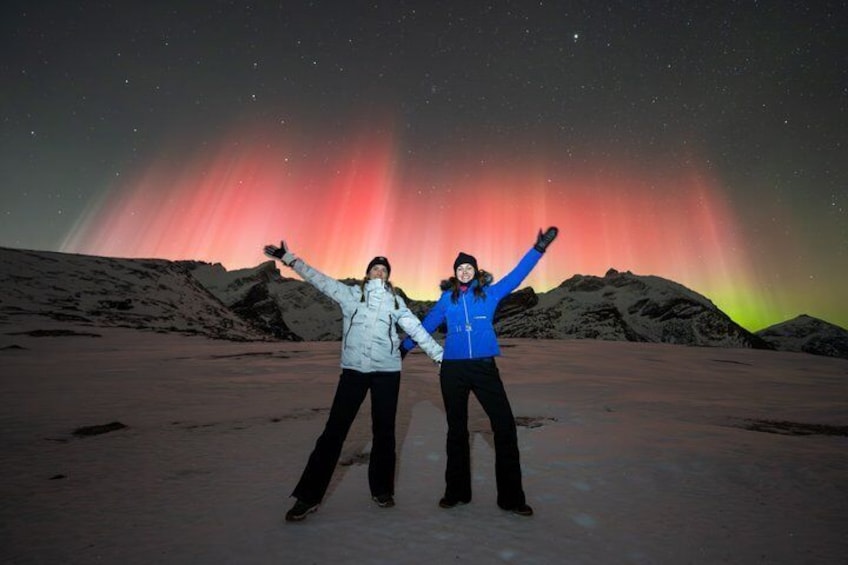 Two people standing on a flat surface with a colorful aurora display in the background. 5